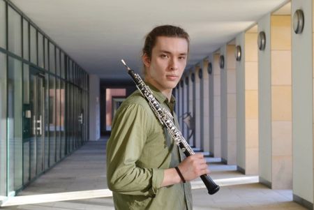 Foto von Leonid Surkov – Halbtotale des Oboisten mit seiner Oboe über der Schulter, draussen in einem Eingangsbereich mit Glastüren, im grünen Hemd, blickt direkt in die Kamera
