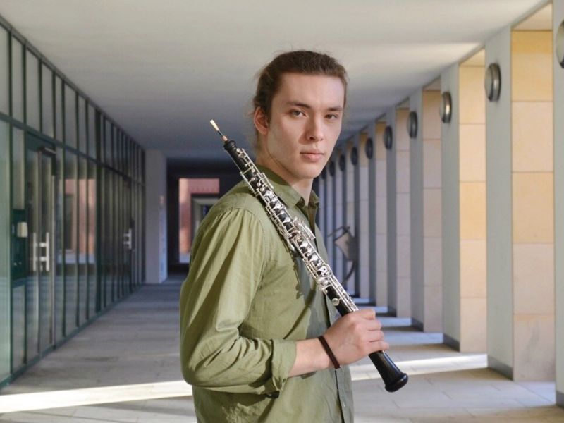 Foto von Leonid Surkov – Halbtotale des Oboisten mit seiner Oboe über der Schulter, draussen in einem Eingangsbereich mit Glastüren, im grünen Hemd, blickt direkt in die Kamera