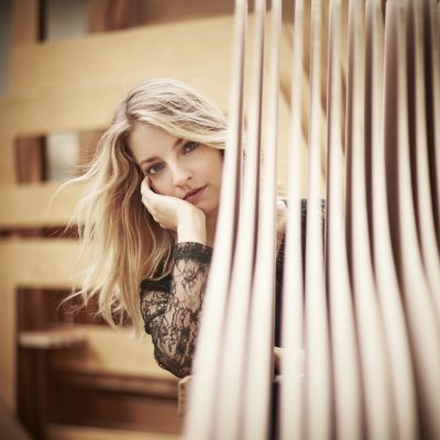 Foto von Lise de la Salle – die Pianistin sieht hinter gestapelten Holzrahmen in einer Klavierfabrik hervor mit der Hand an der Wange
