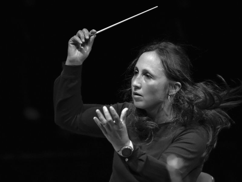 Foto von Joana Carneiro – Halbtotale der Dirigentin, die gerade im schwarzen Hemd mitten in einem Konzert mit Taktstock in der Hand dirigiert
