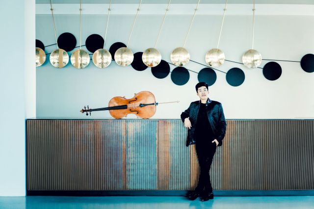 Foto von Bryan Cheng – Totale des Cellisten, angelehnt an einer langen Kommode, dort liegt sein Cello drauf