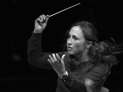 Foto von Joana Carneiro – Halbtotale der Dirigentin, die gerade im schwarzen Hemd mitten in einem Konzert mit Taktstock in der Hand dirigiert