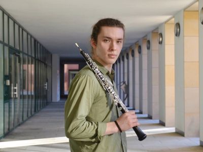 Foto von Leonid Surkov – Halbtotale des Oboisten mit seiner Oboe über der Schulter, draussen in einem Eingangsbereich mit Glastüren, im grünen Hemd, blickt direkt in die Kamera