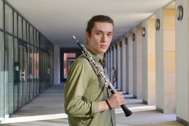 Foto von Leonid Surkov – Halbtotale des Oboisten mit seiner Oboe über der Schulter, draussen in einem Eingangsbereich mit Glastüren, im grünen Hemd, blickt direkt in die Kamera